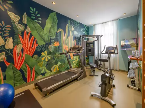 Ein modernes Fitnessstudio mit einem Laufband, einem Heimtrainer und einem Multigym. Die Wände sind mit bunten Pflanzendekoren gestaltet. Ein blauer Gymnastikball liegt auf dem Boden. Natürliches Licht fällt durch ein Fenster mit Vorhängen.