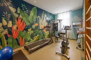 Ein modernes Fitnessstudio mit einem Laufband, einem Heimtrainer und einem Multigym. Die Wände sind mit bunten Pflanzendekoren gestaltet. Ein blauer Gymnastikball liegt auf dem Boden. Natürliches Licht fällt durch ein Fenster mit Vorhängen.