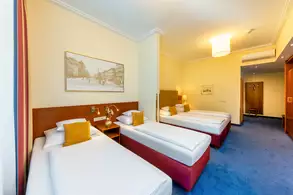 Ein helles Hotelzimmer mit zwei Einzelbetten und einem Doppelbett. An der Wand hängt ein Bild einer Stadtansicht. Es gibt einen Tisch mit einer Lampe und eine Tür, die zu einem weiteren Raum führt. Der Boden ist mit blauem Teppich ausgelegt.