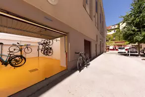 Ein Fahrradabstellraum mit mehreren Fahrrädern an Wänden aufgehängt. Der Boden ist in einem leuchtenden Gelb gestrichen. Rechts davon steht ein weiteres Fahrrad auf dem Bürgersteig. Im Hintergrund ist ein Teil eines mehrstöckigen Gebäudes und einige Autos zu sehen.