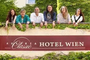 Sechs Personen stehen lächelnd an einem Geländer, das mit Efeu bewachsen ist. Auf einem Schild steht "Austria Classic Hotel Wien". Der Hintergrund besteht aus grünen Pflanzen und einem Gebäude. Die Gruppenmitglieder scheinen freundlich und einladend.