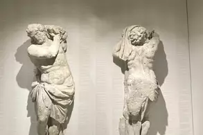 Zwei antike Skulpturen stehen nebeneinander an einer Wand. Die linke Figur zeigt einen Mann, der seine Arme hebt, während die rechte Figur einen Mann zeigt, der sich bekleidet. Beide sind detailliert gearbeitet und aus hellem Material.