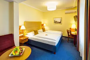 Ein Hotelzimmer mit zwei Einzelbetten, einer Sitzgelegenheit, einem Tisch mit Obstschale und einem Schreibtisch. Es gibt warme Farben, eine angenehme Beleuchtung und ein Fenster mit Vorhängen. Ein Bild an der Wand rundet die Einrichtung ab.