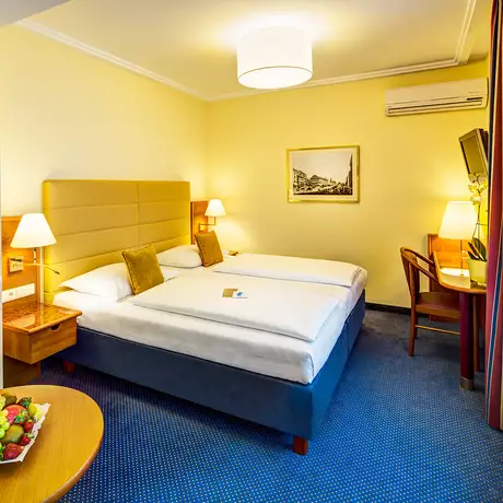 Ein Hotelzimmer mit zwei Einzelbetten, einer Sitzgelegenheit, einem Tisch mit Obstschale und einem Schreibtisch. Es gibt warme Farben, eine angenehme Beleuchtung und ein Fenster mit Vorhängen. Ein Bild an der Wand rundet die Einrichtung ab.