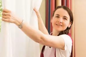 Eine Frau mit braunen Haaren zieht einen weißen Vorhang an einem Fenster auf. Sie trägt ein T-Shirt und eine Schürze. Im Hintergrund sind bunte Streifen an der Wand zu sehen. Die Gesichtsausdruck der Frau ist freundlich und konzentriert.