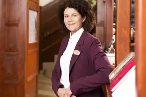 Eine lächelnde Frau steht im Eingangsbereich eines Gebäudes. Sie trägt eine dunkle Jacke und ein helles Blusenshirt. Im Hintergrund ist eine Treppe und einige Holzelemente sichtbar. Die Szene vermittelt eine einladende Atmosphäre.