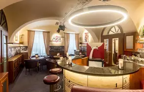 Das Bild zeigt eine elegante Bar mit einem runden Lichtkranz, stilvollen Möbeln und einem ansprechend gestalteten Innenraum. Fenster mit Vorhängen und eine dezente Dekoration verleihen dem Raum eine gemütliche Atmosphäre. Es sind Tische und Stühle vorhanden, die zum Verweilen einladen.