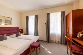 Ein helles Hotelzimmer mit einem Doppelbett, einem kleinen roten Hocker, einem Tisch mit einem Stuhl und einem Kleiderschrank. Große Fenster mit Vorhängen lassen Tageslicht herein. An der Wand hängt ein gerahmtes Bild.