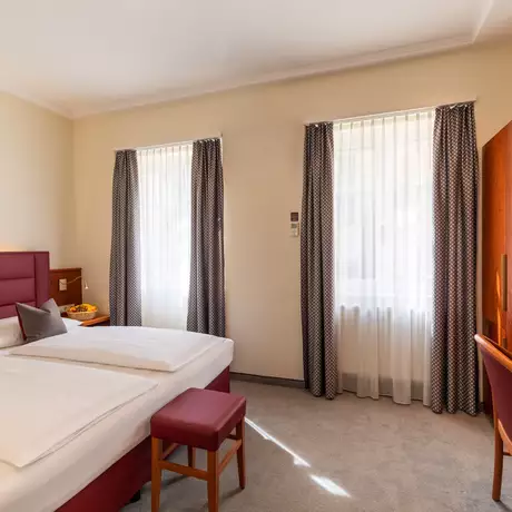 Ein helles Hotelzimmer mit einem Doppelbett, einem kleinen roten Hocker, einem Tisch mit einem Stuhl und einem Kleiderschrank. Große Fenster mit Vorhängen lassen Tageslicht herein. An der Wand hängt ein gerahmtes Bild.