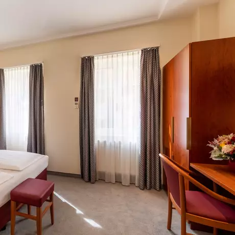 Ein helles Hotelzimmer mit einem Doppelbett, einem kleinen roten Hocker, einem Tisch mit einem Stuhl und einem Kleiderschrank. Große Fenster mit Vorhängen lassen Tageslicht herein. An der Wand hängt ein gerahmtes Bild.
