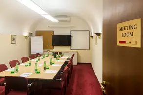 Ein Konferenzraum mit einem langen Tisch, umgeben von Stühlen. Auf dem Tisch stehen grüne Glasflaschen. An der Wand befindet sich ein Whiteboard und ein Bildschirm. Die Tür hat ein Schild mit der Aufschrift "MEETING ROOM". Der Raum hat einen roten Teppich und eine helle Beleuchtung.
