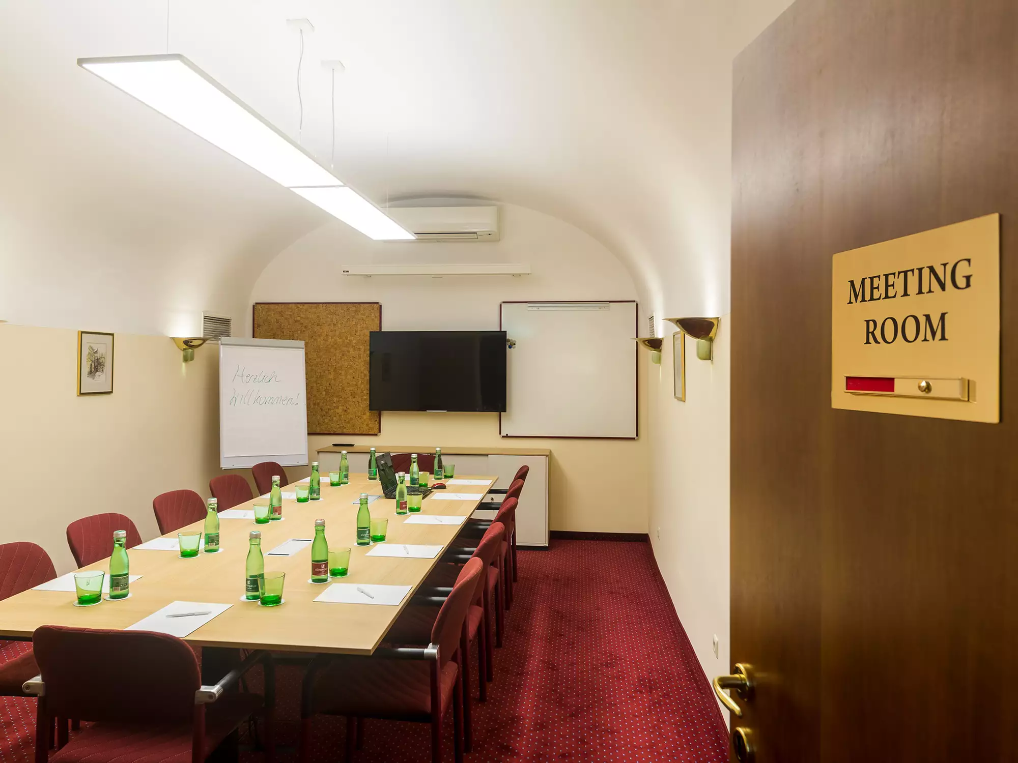 Ein Konferenzraum mit einem langen Tisch, umgeben von Stühlen. Auf dem Tisch stehen grüne Glasflaschen. An der Wand befindet sich ein Whiteboard und ein Bildschirm. Die Tür hat ein Schild mit der Aufschrift "MEETING ROOM". Der Raum hat einen roten Teppich und eine helle Beleuchtung.