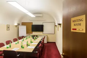 Ein Konferenzraum mit einem langen Tisch, umgeben von Stühlen. Auf dem Tisch stehen grüne Glasflaschen. An der Wand befindet sich ein Whiteboard und ein Bildschirm. Die Tür hat ein Schild mit der Aufschrift "MEETING ROOM". Der Raum hat einen roten Teppich und eine helle Beleuchtung.
