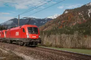 Ein roter Zug der ÖBB fährt entlang einer Gleisanlage, umgeben von einer herbstlichen Landschaft mit Bergen im Hintergrund und teilweise bewaldeten Flächen. Der Himmel ist klar mit vereinzelten Wolken.