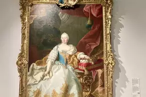 Ein opulentes Gemälde einer königlichen Frau in einem prunkvollen Kleid mit goldenen Verzierungen. Sie sitzt an einem Tisch, auf dem eine Torte steht. Der Rahmen ist kunstvoll verziert und zeigt oben ein kleines Bildnis.