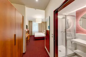 Ein helles Hotelzimmer mit einem angrenzenden Badezimmer. Auf der linken Seite befindet sich ein großer Kleiderschrank, während rechts ein modernes Bad mit Dusche und Waschbecken zu sehen ist. Im Hintergrund steht ein Doppelbett, umgeben von Vorhängen und einem Fernseher.