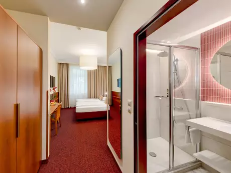 Ein helles Hotelzimmer mit einem angrenzenden Badezimmer. Auf der linken Seite befindet sich ein großer Kleiderschrank, während rechts ein modernes Bad mit Dusche und Waschbecken zu sehen ist. Im Hintergrund steht ein Doppelbett, umgeben von Vorhängen und einem Fernseher.