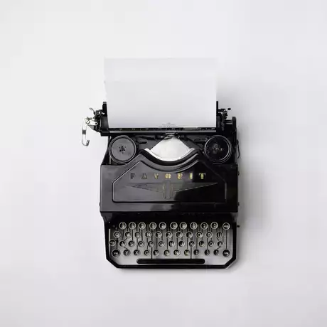 Eine schwarze Schreibmaschine von oben mit einem leeren Blatt Papier, das aus dem Gerät herausragt. Die Tasten der Maschine sind gut sichtbar. Der Hintergrund ist einfarbig hell.