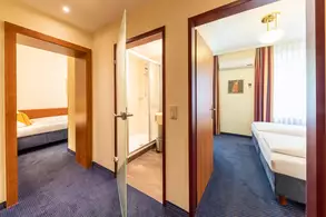 Ein Blick in einen Hotelflur mit zwei offenen Türen. Die linke Tür führt zu einem Schlafzimmer mit einem Bett, die rechte Tür öffnet sich zu einem Badezimmer mit Dusche. Der Boden ist mit blauen Teppich ausgelegt, und Licht strömt durch ein Fenster in den Raum.