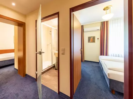 Ein Blick in einen Hotelflur mit zwei offenen Türen. Die linke Tür führt zu einem Schlafzimmer mit einem Bett, die rechte Tür öffnet sich zu einem Badezimmer mit Dusche. Der Boden ist mit blauen Teppich ausgelegt, und Licht strömt durch ein Fenster in den Raum.