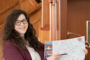 Eine Person mit lockigem Haar und Brille lächelt und hält eine Karte in der Hand. Sie steht in einer stilvollen Umgebung mit Holzvertäfelung im Hintergrund und zeigt mit einem Stift auf die Karte.